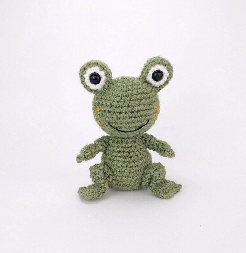 May include: A green crocheted frog toy with white eyes and a black smile. The frog is sitting on a white background.