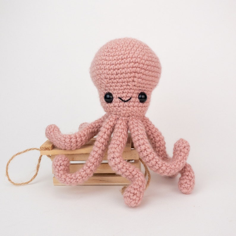 May include: A pink crocheted octopus with black eyes and a smiling mouth. The octopus is sitting on a small wooden crate.