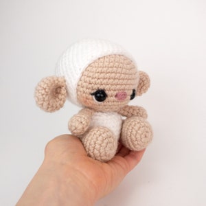 May include: A small crocheted lamb toy with a white hat, brown body, and black eyes. The lamb is sitting on a hand.