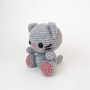 Puede incluir: Un gato de crochet gris con almohadillas de patas rosas y ojos negros. El gato est&aacute; sentado con la cola enroscada alrededor de sus patas.