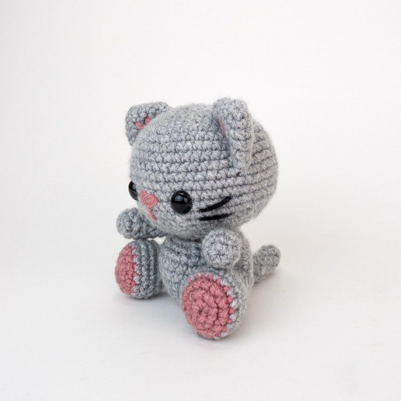 Puede incluir: Un gato de crochet gris con almohadillas de patas rosas y ojos negros. El gato est&aacute; sentado con la cola enroscada alrededor de sus patas.
