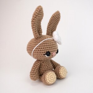 May include: A brown crocheted bunny with white ears and a white flower headband. The bunny has black eyes and a pink nose.
