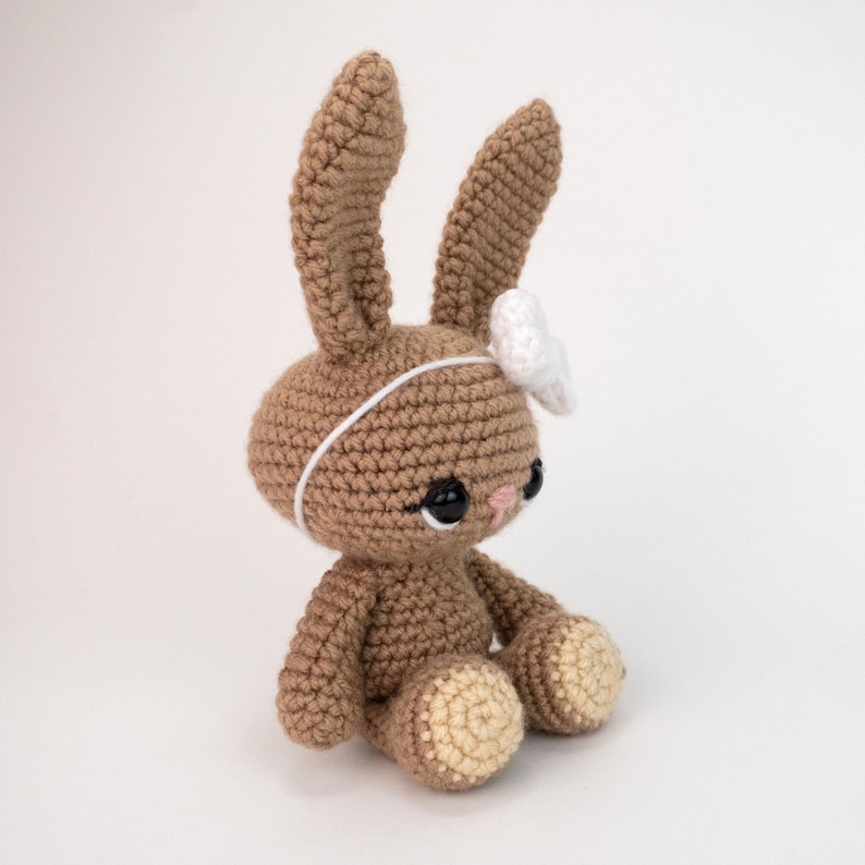May include: A brown crocheted bunny with white ears and a white flower headband. The bunny has black eyes and a pink nose.