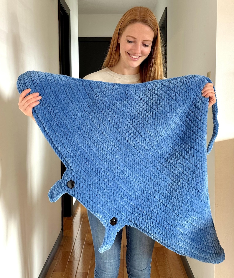 May include: A blue crocheted manta ray blanket with black eyes. The blanket is being held by a person.