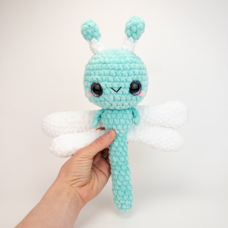 May include: A crocheted dragonfly toy with a light blue body, white wings, and large black eyes. The dragonfly has a small, stitched smile.