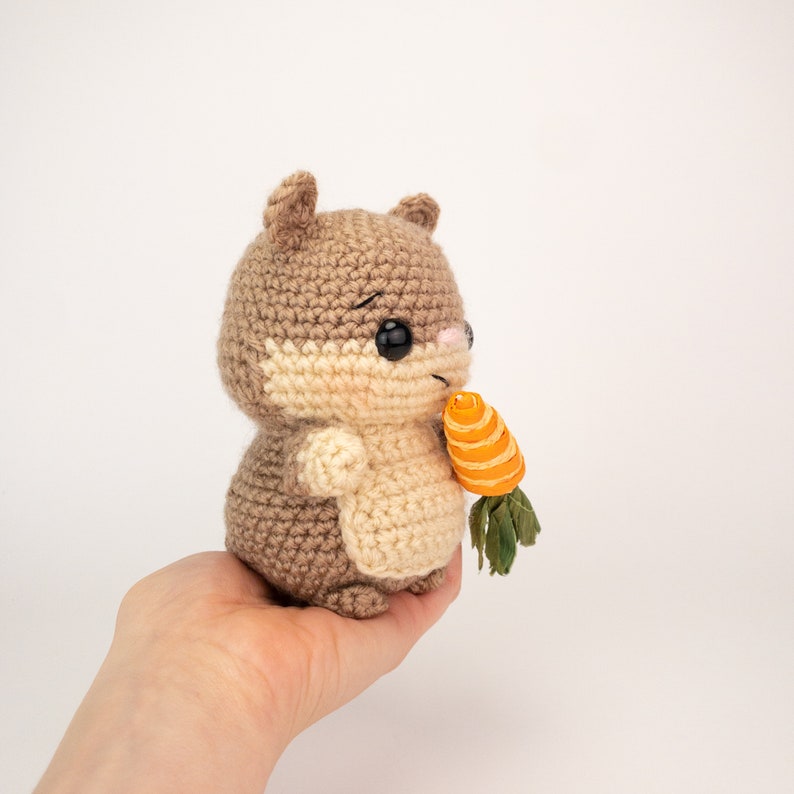 May include: A crocheted brown and beige squirrel holding a small orange carrot with green leaves.