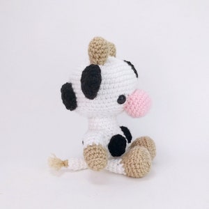 Puede incluir: Un juguete de peluche de vaca tejido a crochet con manchas blancas y negras, orejas marrones y una nariz rosa. La vaca est&aacute; sentada con las piernas cruzadas y tiene una cola peque&ntilde;a.