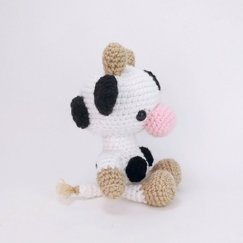 Puede incluir: Un juguete de peluche de vaca tejido a crochet con manchas blancas y negras, orejas marrones y una nariz rosa. La vaca est&aacute; sentada con las piernas cruzadas y tiene una cola peque&ntilde;a.