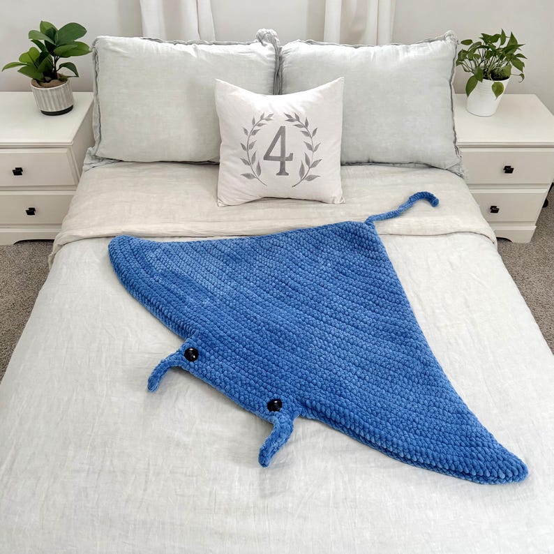 May include: A blue crocheted manta ray blanket with black eyes. The blanket is on a white bed with a pillow that says "4" in a wreath.