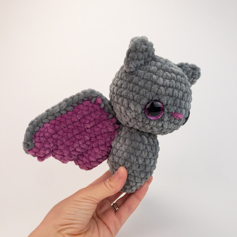 K&ouml;nnte beinhalten: Eine graue geh&auml;kelte Fledermaus mit rosa Fl&uuml;geln und gro&szlig;en lila Augen. Die Fledermaus wird in einer Hand gehalten.