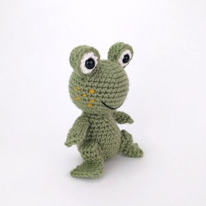 May include: A green crocheted frog toy with white eyes and yellow stitching. The frog is standing on a white surface.