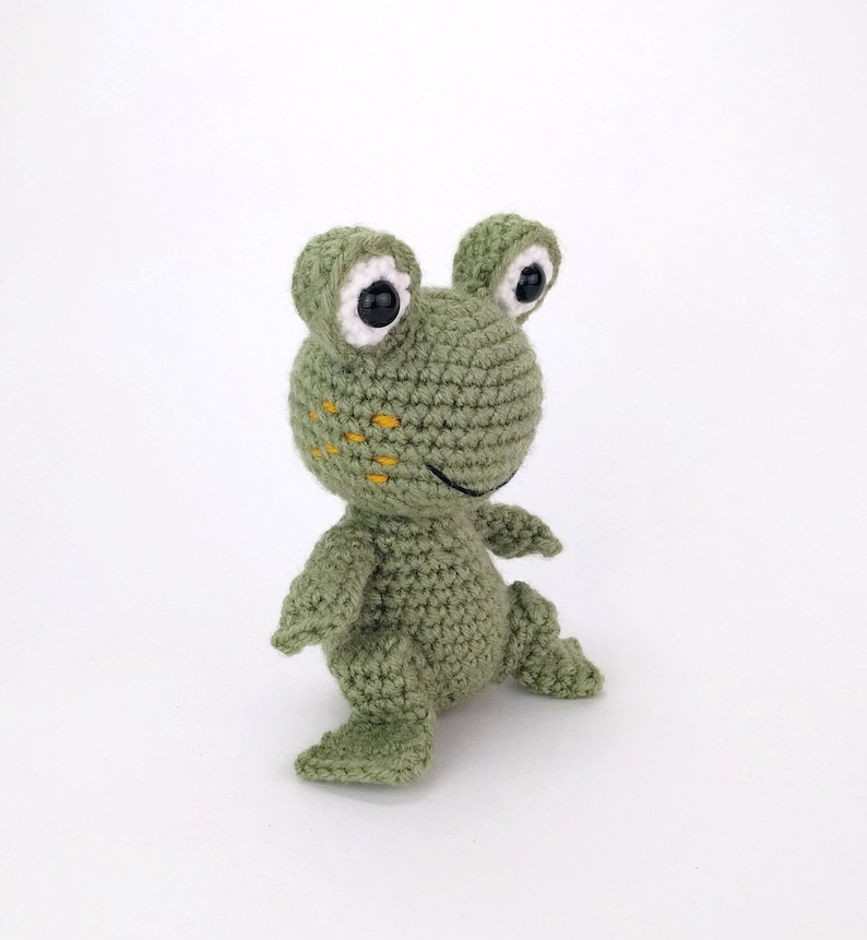 May include: A green crocheted frog toy with white eyes and yellow stitching. The frog is standing on a white surface.