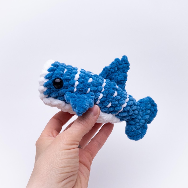May include: A blue and white crocheted whale plush toy with black eyes. The whale is being held in a hand.