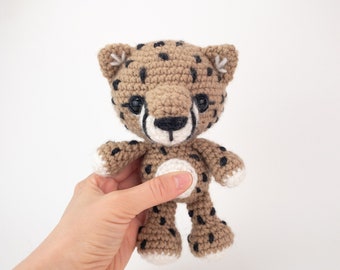 May include: A crocheted cheetah toy with brown and black fur, white paws, and black eyes. The toy is being held by a hand.