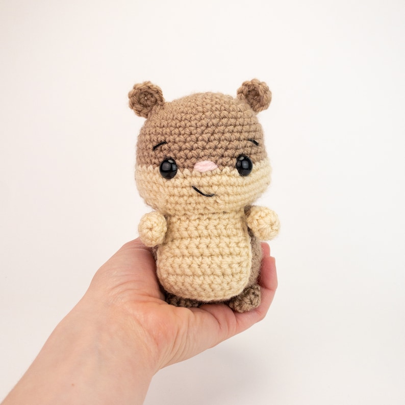 May include: A crocheted brown and beige squirrel plush toy with black eyes and a pink nose. The toy is being held in a person's hand.