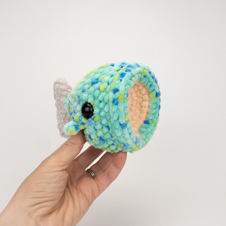 May include: A crocheted plush toy shaped like a fish with a light blue, green, and yellow speckled body and a light gray tail fin. The toy has a black button eye.