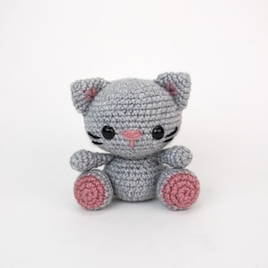 Puede incluir: Un juguete de crochet de gato gris con almohadillas de patas rosas y ojos negros. El gato tiene una nariz rosa y bigotes negros.