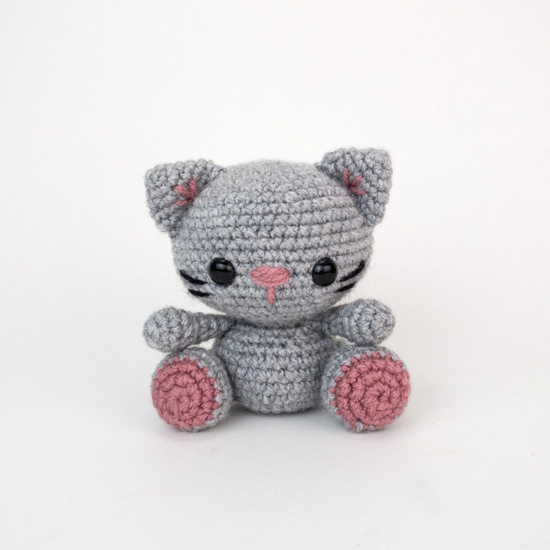 Puede incluir: Un juguete de crochet de gato gris con almohadillas de patas rosas y ojos negros. El gato tiene una nariz rosa y bigotes negros.