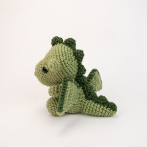 May include: A green crocheted dragon with a small, cute face and wings. The dragon is sitting on a white background.