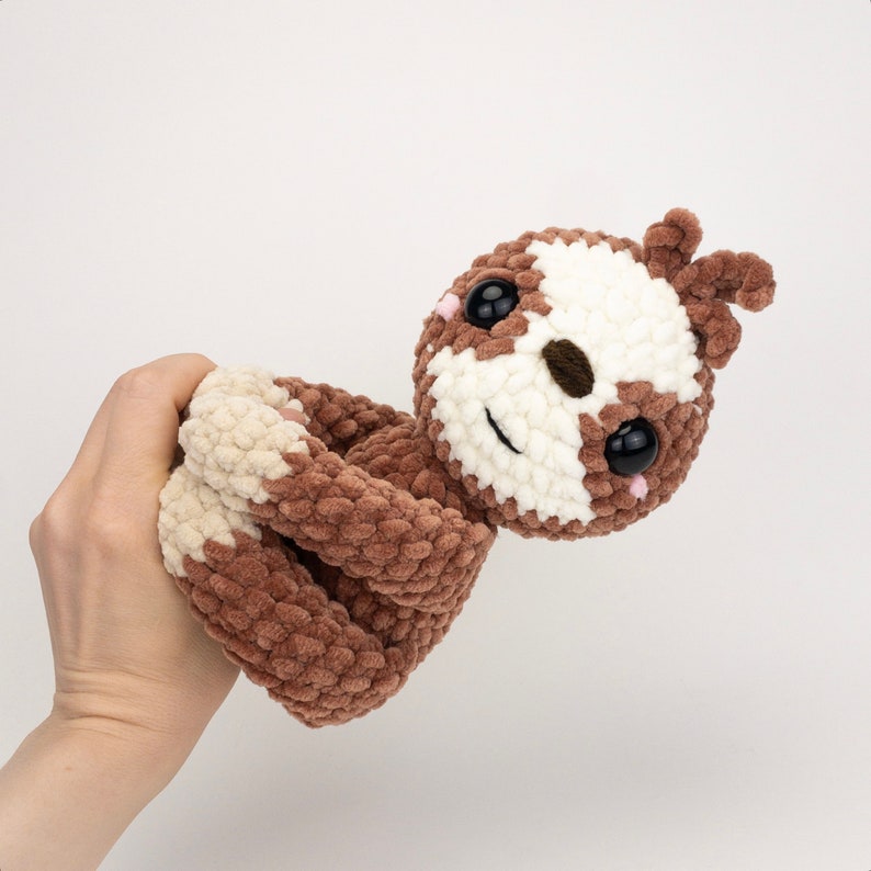 May include: A brown and white crocheted sloth toy with large black eyes and a pink nose. The sloth is curled up in a relaxed pose.