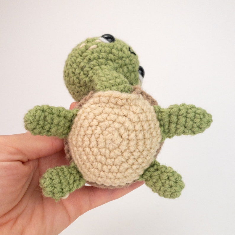 May include: A crocheted turtle toy with a green shell and beige underside. The turtle has black eyes and a small, crocheted mouth.