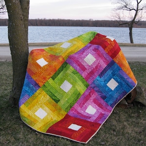 Op de afbeelding: Een kleurrijk quilt met een patchwork-ontwerp met vierkanten in rood, oranje, geel, groen, blauw en paars. De quilt is gemaakt van verschillende stoffen en heeft een witte achtergrond.
