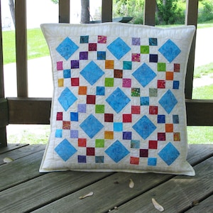 May include: A white pillow with a blue and white diamond pattern. The pillow is made of fabric squares sewn together in a patchwork design. The pillow is sitting on a wooden surface.