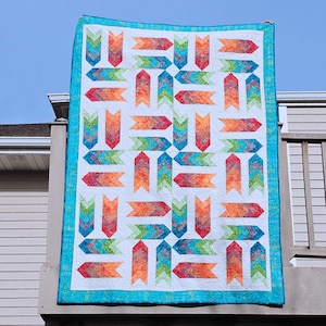 May include: A rectangular quilt featuring a repeating pattern of chevron-style arrows in a vibrant rainbow gradient of red, orange, yellow, green, and blue. The quilt has a white background and is finished with a solid bright turquoise border.