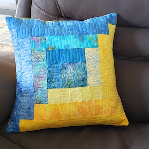 Może przedstawiać: Poduszka patchworkowa w kolorze żółtym i niebieskim z kwadratowym wzorem. Poduszka jest wykonana z różnych tkanin i ma pikowaną fakturę.