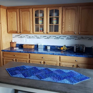May include: A blue quilted table runner with a geometric pattern. The runner is laid out on a wooden table in front of a kitchen with wooden cabinets and a blue countertop.