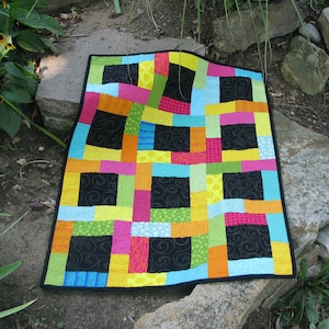 May include: A colourful patchwork quilt with a geometric design. The quilt is made up of squares and rectangles in various shades of pink, yellow, green, blue, orange, and black.