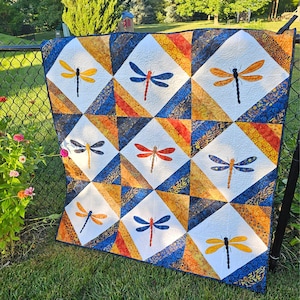 May include: A handmade quilt featuring a repeating pattern of dragonflies. Each dragonfly is a different color, set against a white square. The quilt has a blue, orange, and gold border, and is displayed outdoors on a fence.