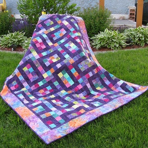 May include: A vibrant quilt with a geometric pattern, set against a dark purple backdrop. It incorporates squares of diverse colours such as pink, blue, and orange. The quilt is displayed on green grass.