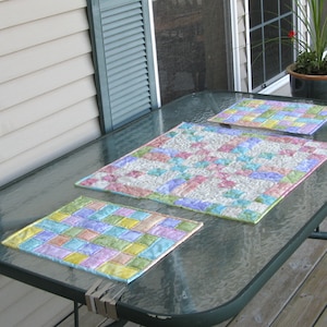 May include: Three pastel-colored quilted placemats on a glass table top. The placemats are made of a patchwork of squares in shades of pink, green, yellow, and blue.