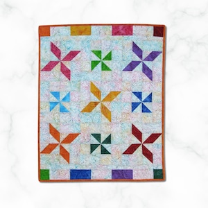 Könnte beinhalten: Ein farbenfrohes Quilt mit einem Windrad-Design. Das Quilt zeigt eine Vielzahl von leuchtenden Farben, darunter Rosa, Grün, Lila, Blau, Orange und Rot. Die Windräder sind vor einem hellen Hintergrund mit einem floralen Muster angeordnet, und das Quilt ist mit einem einfarbigen orangefarbenen Stoff eingefasst.