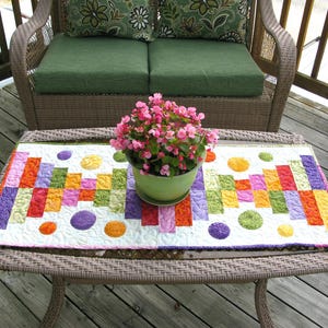 May include: A colourful patchwork table runner with a geometric design in shades of purple, orange, yellow, green, and pink. The runner is placed on a wicker table with a potted plant in the centre.
