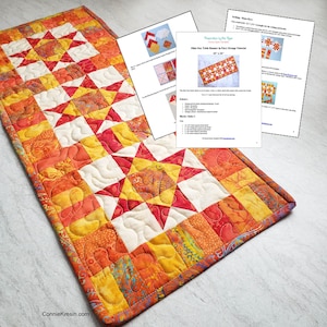 May include: A table runner made with a patchwork of orange, red, and yellow fabric. The runner features a star pattern and is quilted with a wavy design. The runner is 18" x 36" and is titled "Ohio Star Table Runner in Fiery Orange Tutorial".