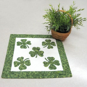 May include: Green shamrock table runner with a white center featuring five shamrock cutouts. The table runner has a green border with a shamrock pattern.