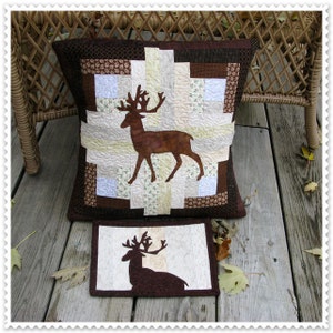 May include: A decorative pillow and matching placemat featuring a deer silhouette. The pillow has a patchwork design with brown, cream, and floral fabrics. The deer is a brown silhouette. The placemat has a similar deer design.