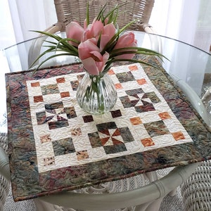 Pode incluir: Um corredor de mesa acolchoado com um padrão floral verde, marrom e laranja. O corredor de mesa está colocado em uma mesa de vidro com um vaso de tulipas rosa no centro.