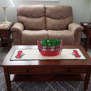May include: A brown wooden coffee table with a white and green runner featuring red trucks and Christmas trees. A red wicker basket with green Christmas trees sits on the runner.