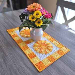 May include: A handmade, quilted table runner featuring a sunflower design. The runner has a patchwork border in shades of yellow and orange, with a white center panel showcasing a detailed sunflower applique. A vase of colorful flowers sits atop the runner.