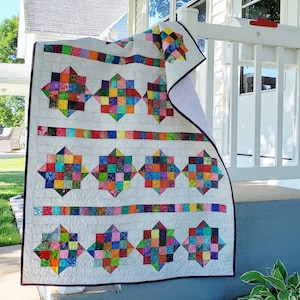May include: A handmade quilt with a white background and colourful star and square patterns. The quilt features vibrant squares of red, blue, green, and yellow, arranged in star shapes and horizontal bands. The quilt is displayed outdoors.