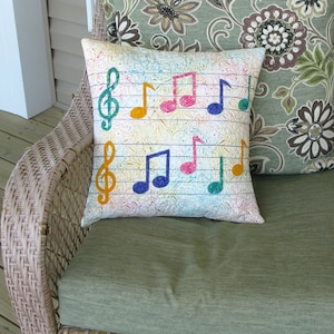 May include: A white pillow with a musical note pattern in various colors, including pink, blue, green, and yellow. The pillow is sitting on a wicker chair with a green cushion.