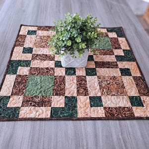 May include: A square, quilted table topper with a patchwork design in shades of brown, green, and beige. A small, white, textured pot with a green plant sits in the center. The quilt has a dark brown border.