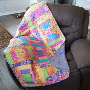 Op de afbeelding: Een kleurrijk patchwork-deken met verschillende bloemen- en vlinderpatronen in tinten roze, geel, groen en blauw. Het deken is gevouwen en over een bruine fauteuil gelegd.