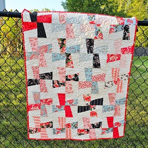 May include: A handmade quilt with a cream background and a patchwork design. The quilt features red, black, pink, and blue floral and patterned fabric squares. The quilt is trimmed with a red border and has quilted stitching.