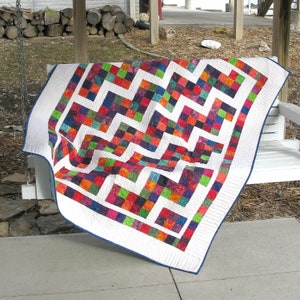 May include: A colorful patchwork quilt with a white background and a zig-zag pattern in white. The quilt is draped over a white wooden bench.