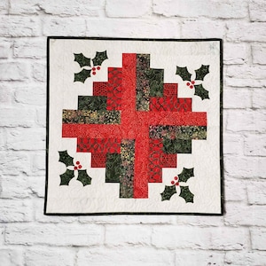May include: A square quilted wall hanging with a Christmas theme. The design features a red cross shape and green and red patterned blocks. Holly leaf and berry appliqués are in each corner. The quilt is bordered with a dark green binding.