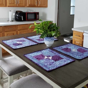 Può includere: Quattro tovagliette all'americana trapuntate viola e blu con un design geometrico bianco sono disposte su un tavolo marrone scuro con una pianta in vaso al centro. Il tavolo è apparecchiato con due sedie bianche.